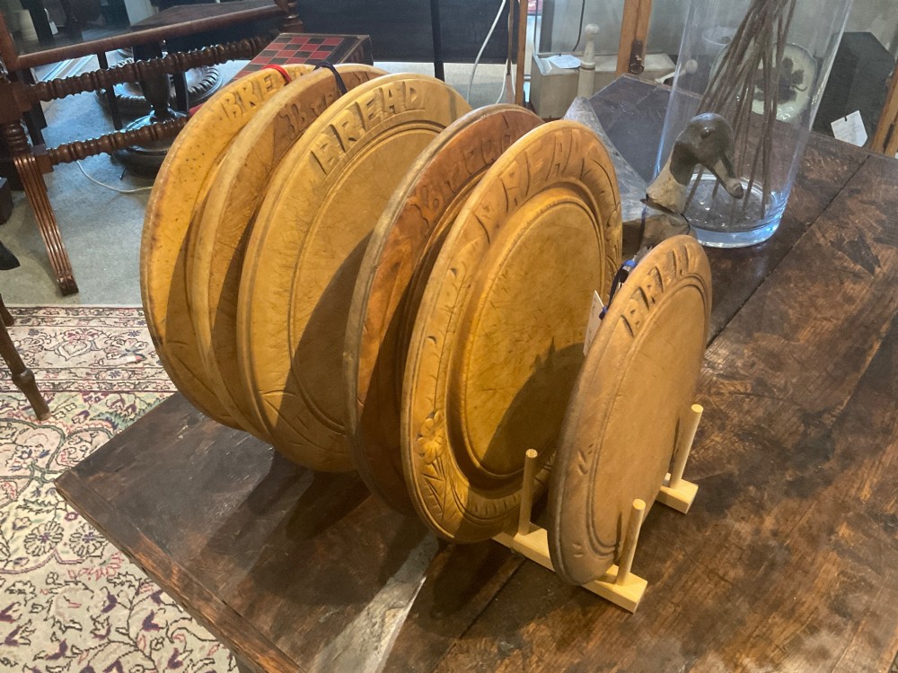 A circular carved wooden bread board with carved lettering spelling BREAD along the outer rim and visible knife marks. View VI