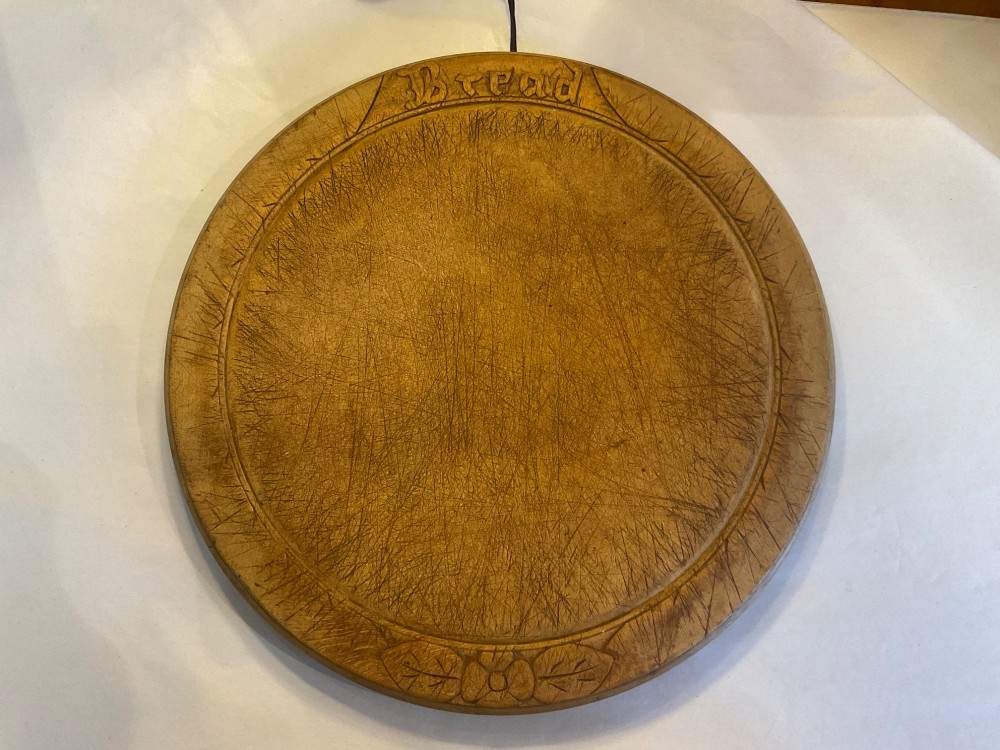 A circular antique carved wooden bread board featuring the word Bread carved into the upper border, leaf motifs carved on the lower border, and central knife marks from usage.