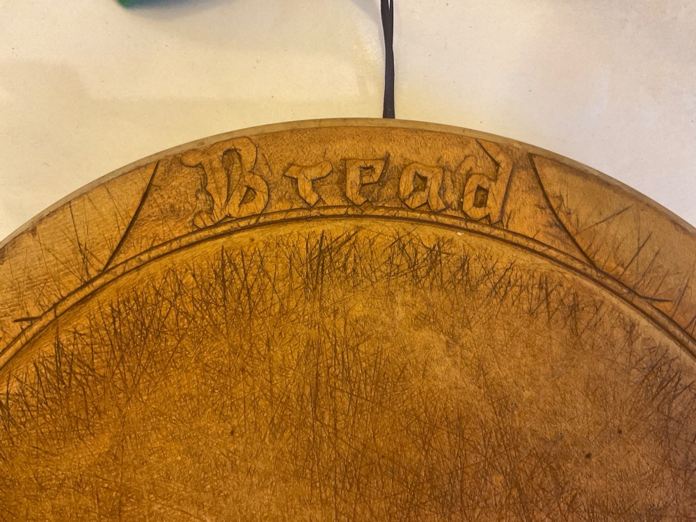 A circular antique carved wooden bread board featuring the word Bread carved into the upper border, leaf motifs carved on the lower border, and central knife marks from usage. View II