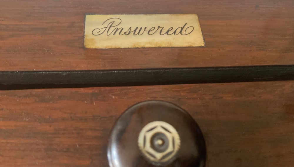 A dark mahogany 19th-century letter box featuring two top slots labelled Answered and Unanswered, a turned central knob, and a front keyhole with an inserted key. View IV