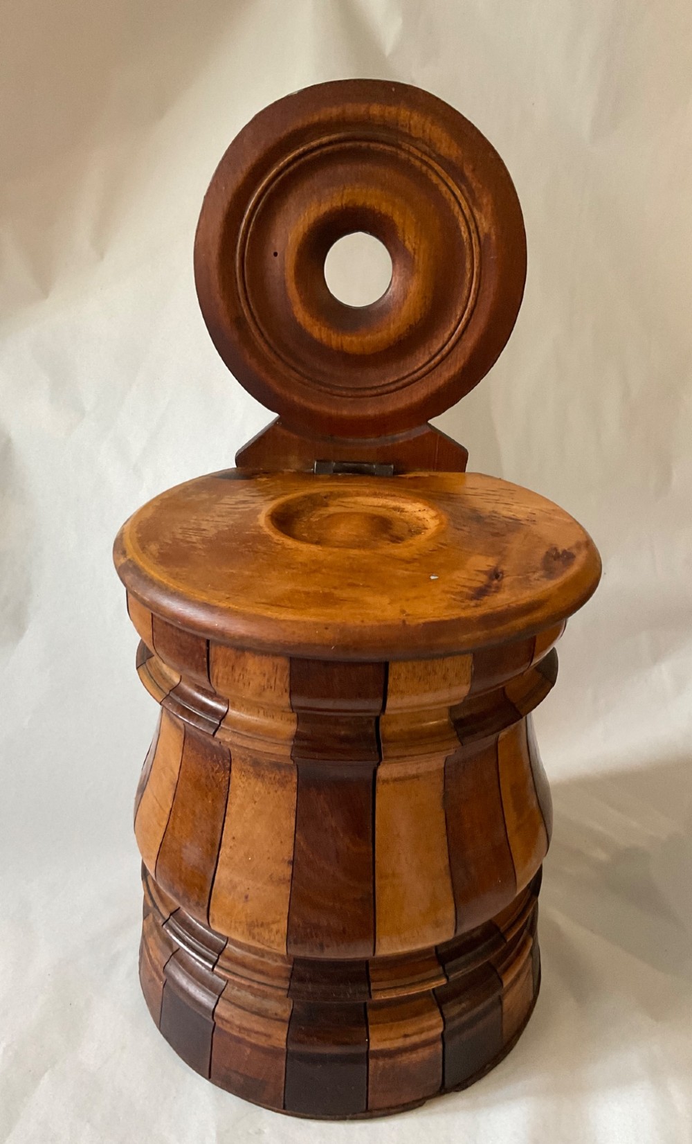 A mid-nineteenth century coopered wooden salt box featuring alternating dark and light timber staves, a circular hinged lid, and an arched backplate with a hanging aperture.