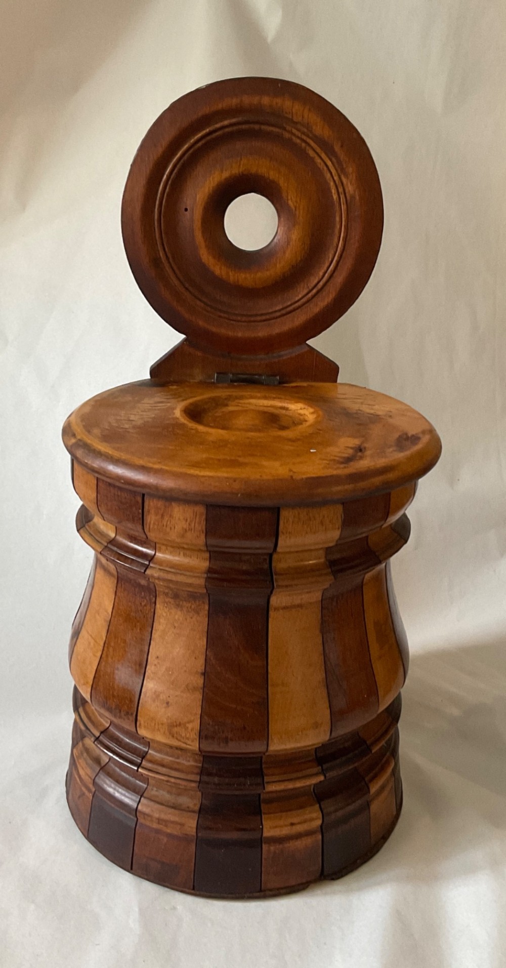 A mid-nineteenth century coopered wooden salt box featuring alternating dark and light timber staves, a circular hinged lid, and an arched backplate with a hanging aperture. View II