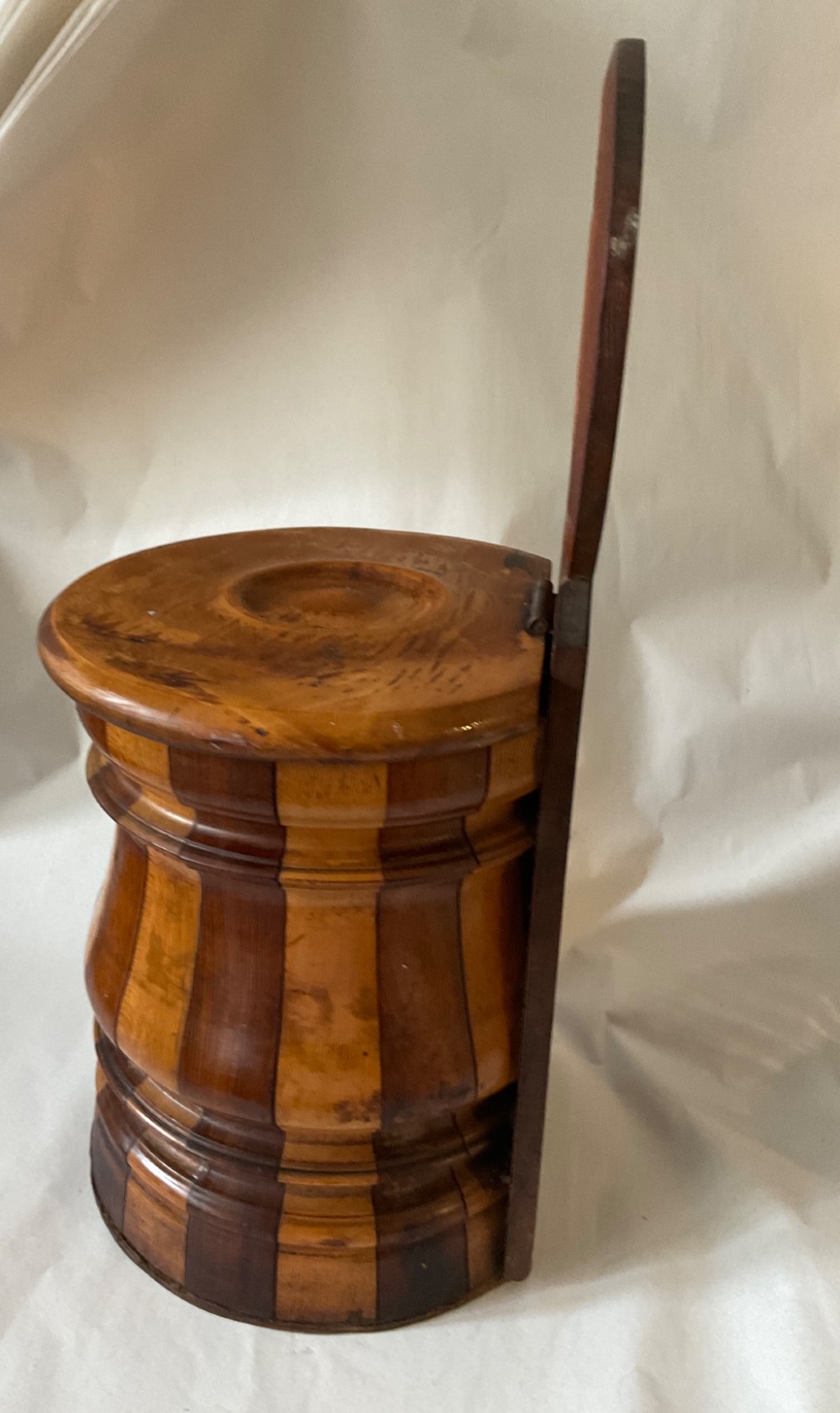 A mid-nineteenth century coopered wooden salt box featuring alternating dark and light timber staves, a circular hinged lid, and an arched backplate with a hanging aperture. View IV
