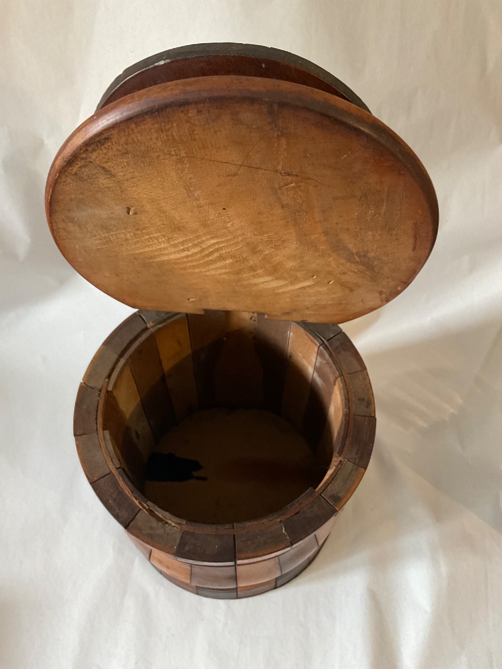 A mid-nineteenth century coopered wooden salt box featuring alternating dark and light timber staves, a circular hinged lid, and an arched backplate with a hanging aperture. View V