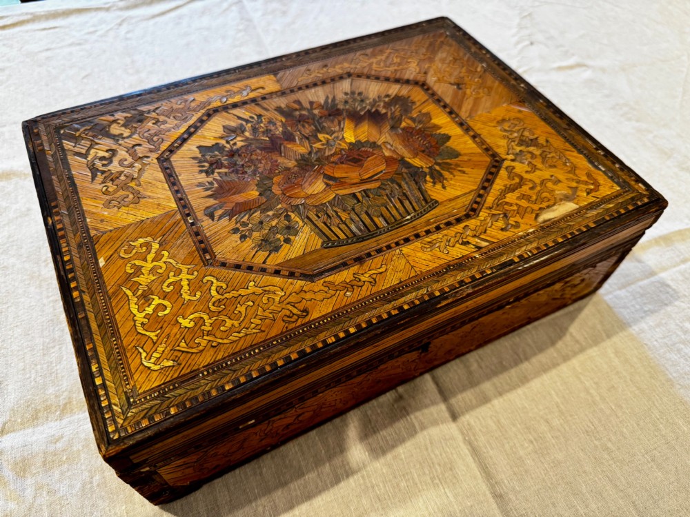 An antique rectangular wooden box embellished with straw marquetry forming a central floral basket surrounded by intricate decorative borders in golden yellow and brown shades.