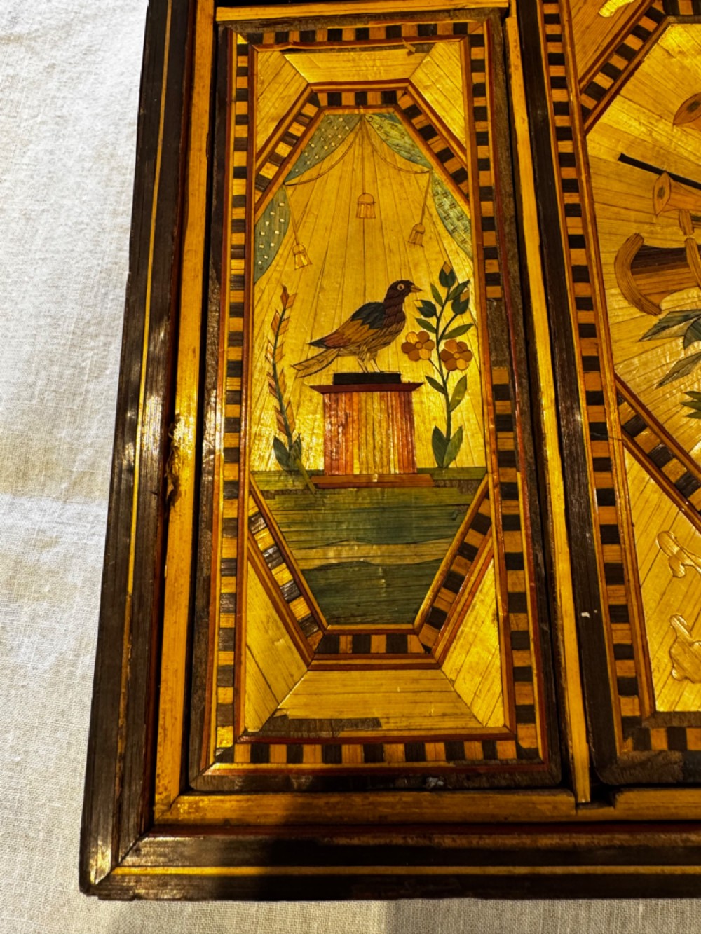 An antique rectangular wooden box embellished with straw marquetry forming a central floral basket surrounded by intricate decorative borders in golden yellow and brown shades. View X