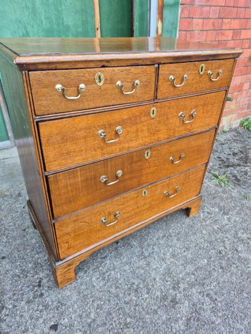 fig 1. A George II golden oak chest of drawers featuring two short drawers over three long graduated drawers with brass swan-neck handles and bracket feet.