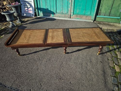 fig 2. An antique campaign-style daybed crafted from dark timber with three woven cane panels, adjustable reclining sections, turned legs, and brass castors.