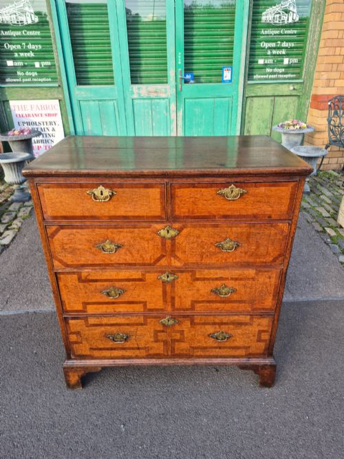 fig 1. An Early Georgian oak and walnut chest of drawers featuring geometric wood banding, engraved brass handles, and bracket feet.