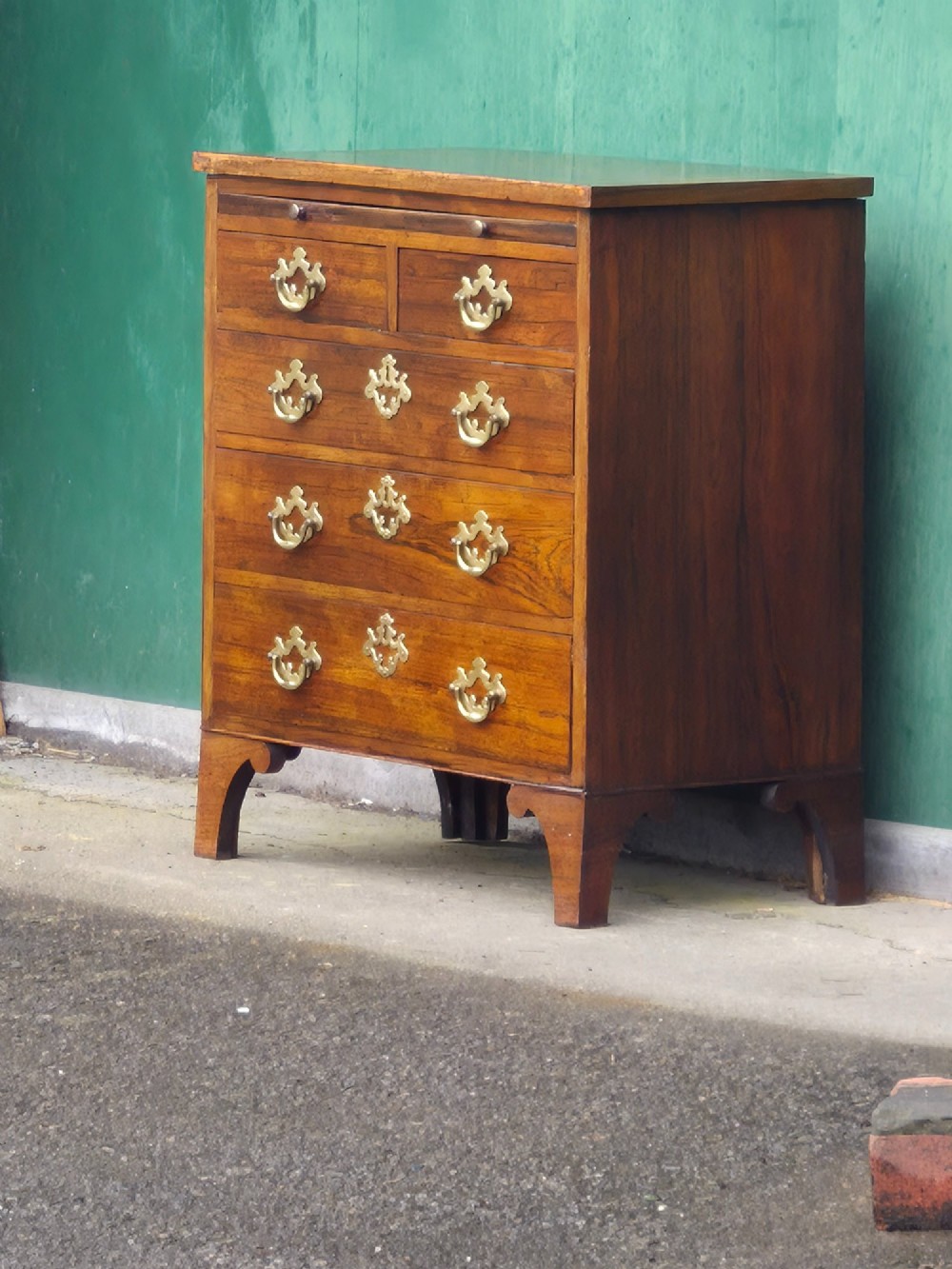 A small rosewood chest of drawers featuring brass handles, bracket feet, and a pull-out brushing slide.
