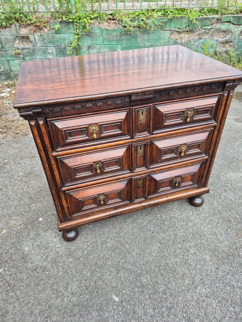 A dark oak chest of drawers featuring three drawers with geometric panel mouldings, brass drop handles, and turned bun feet. View II