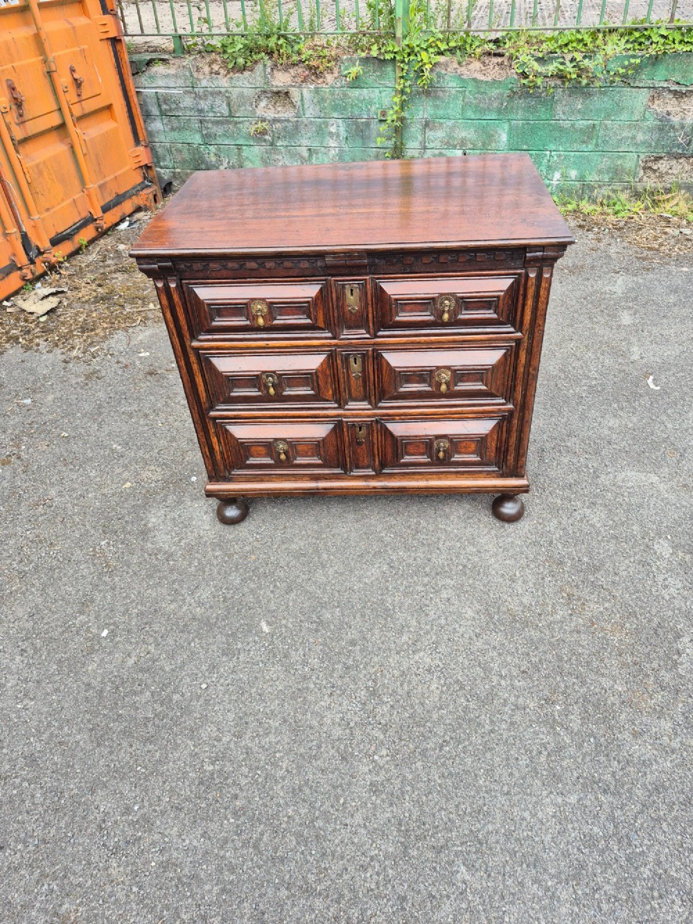 A dark oak chest of drawers featuring three drawers with geometric panel mouldings, brass drop handles, and turned bun feet. View III