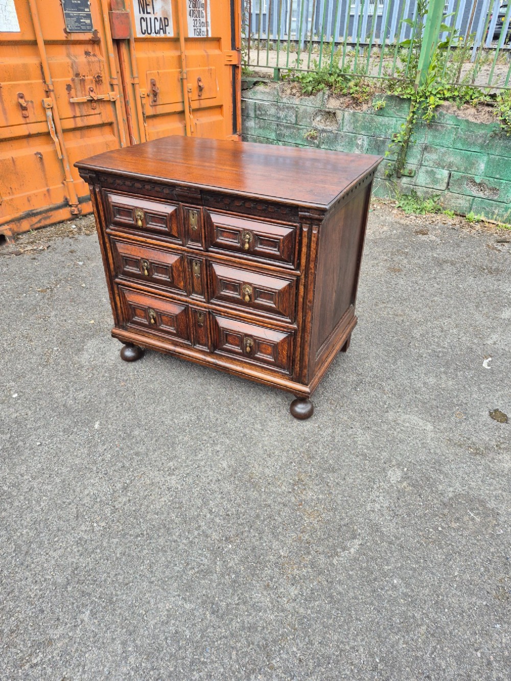 A dark oak chest of drawers featuring three drawers with geometric panel mouldings, brass drop handles, and turned bun feet. View IV