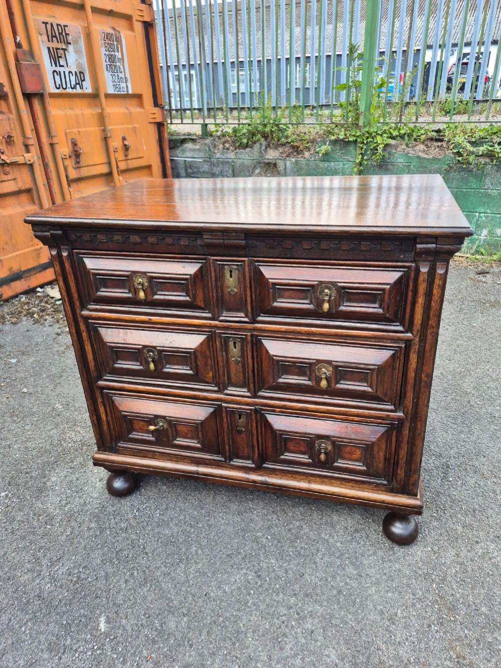 A dark oak chest of drawers featuring three drawers with geometric panel mouldings, brass drop handles, and turned bun feet. View IX