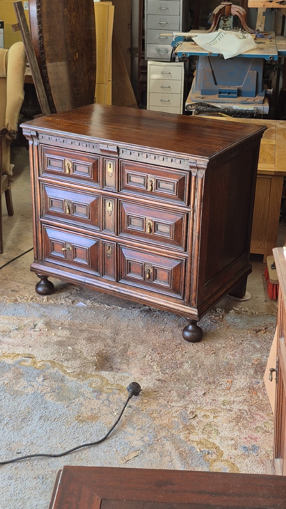 A dark oak chest of drawers featuring three drawers with geometric panel mouldings, brass drop handles, and turned bun feet.