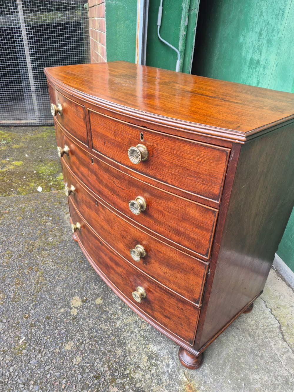 An antique mahogany bowfront chest of drawers featuring two short drawers over three long drawers with brass rosette knobs and turned feet. View III