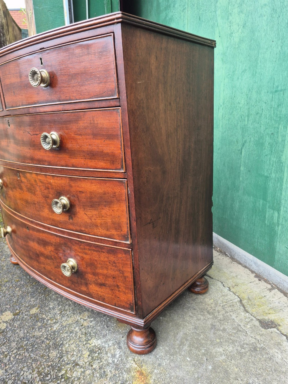 An antique mahogany bowfront chest of drawers featuring two short drawers over three long drawers with brass rosette knobs and turned feet. View VI