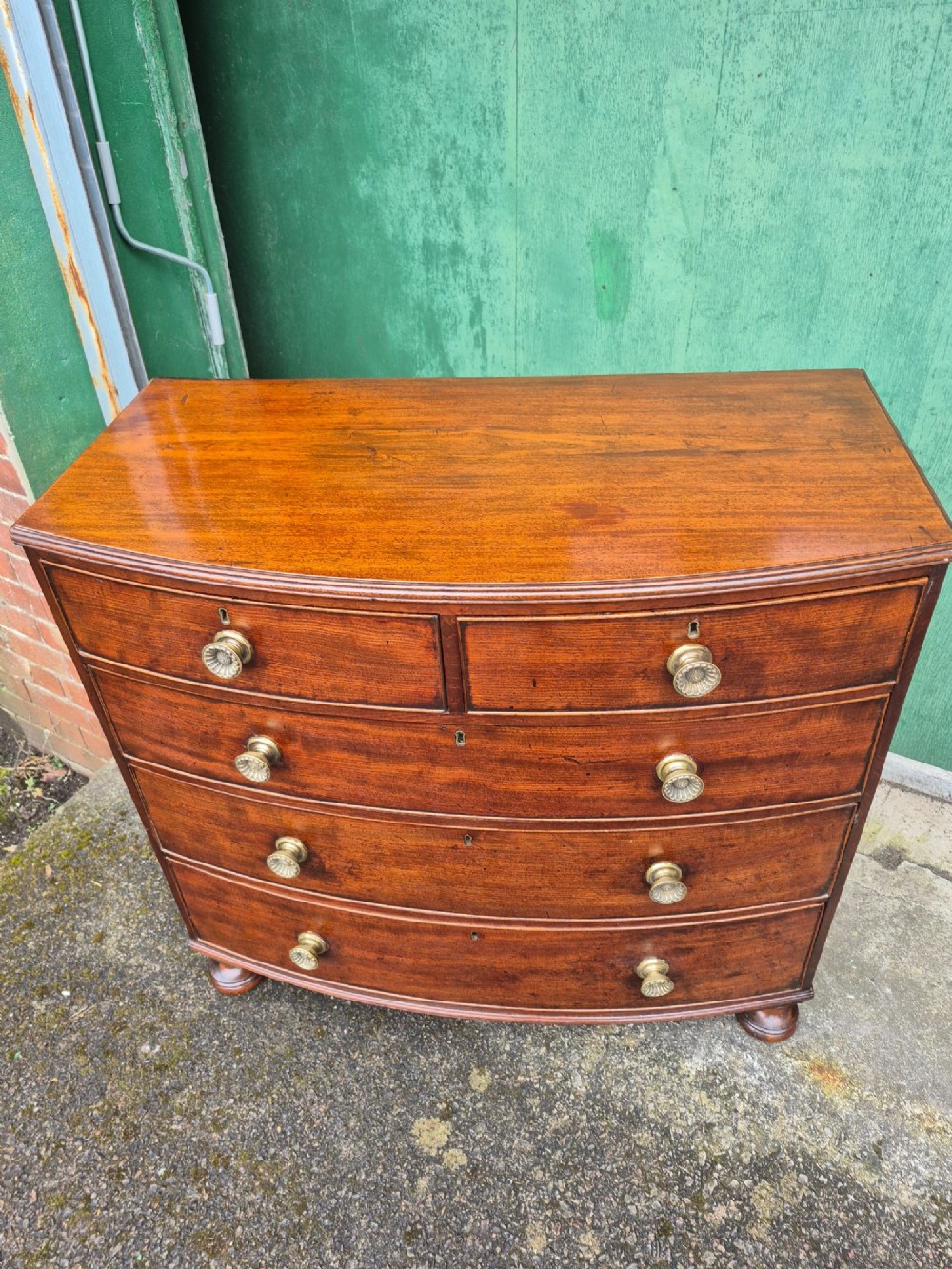 An antique mahogany bowfront chest of drawers featuring two short drawers over three long drawers with brass rosette knobs and turned feet. View VII