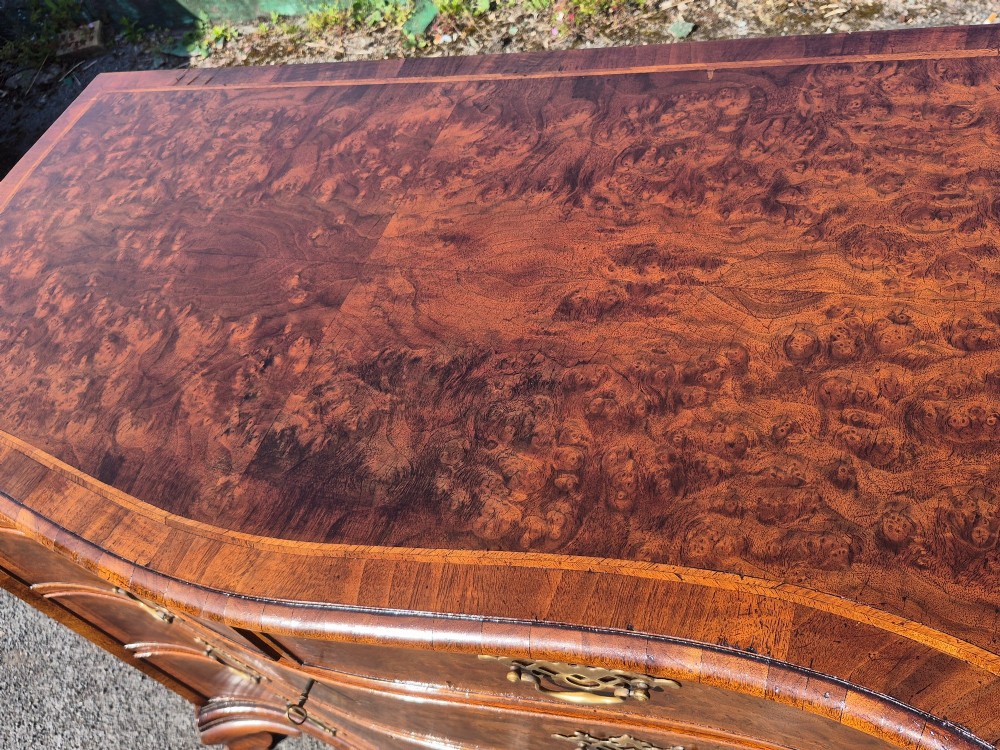 A burr walnut serpentine-front chest of drawers with two short top drawers over three long drawers, featuring brass backplate handles and resting on bracket feet. View V