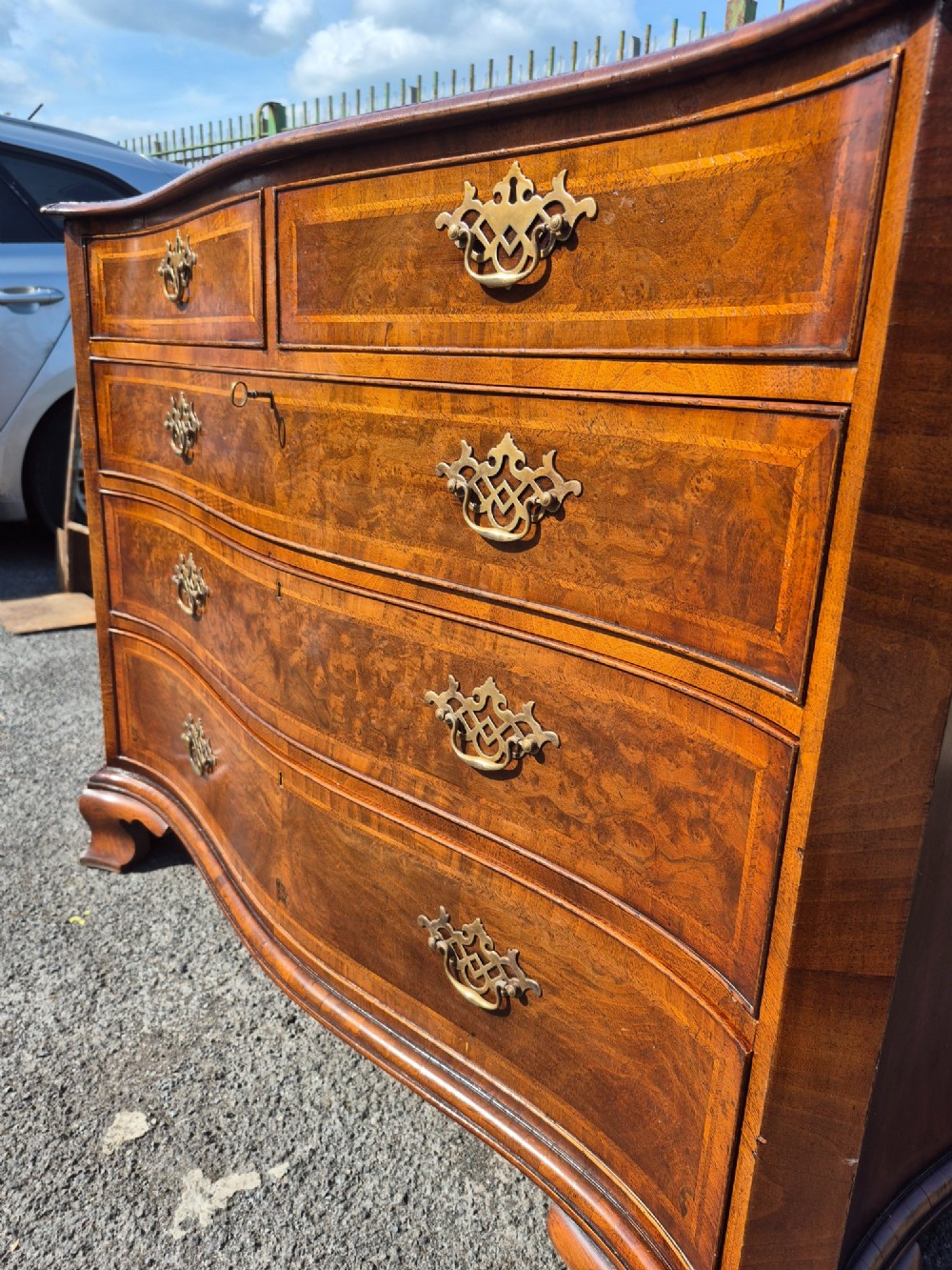 A burr walnut serpentine-front chest of drawers with two short top drawers over three long drawers, featuring brass backplate handles and resting on bracket feet. View X