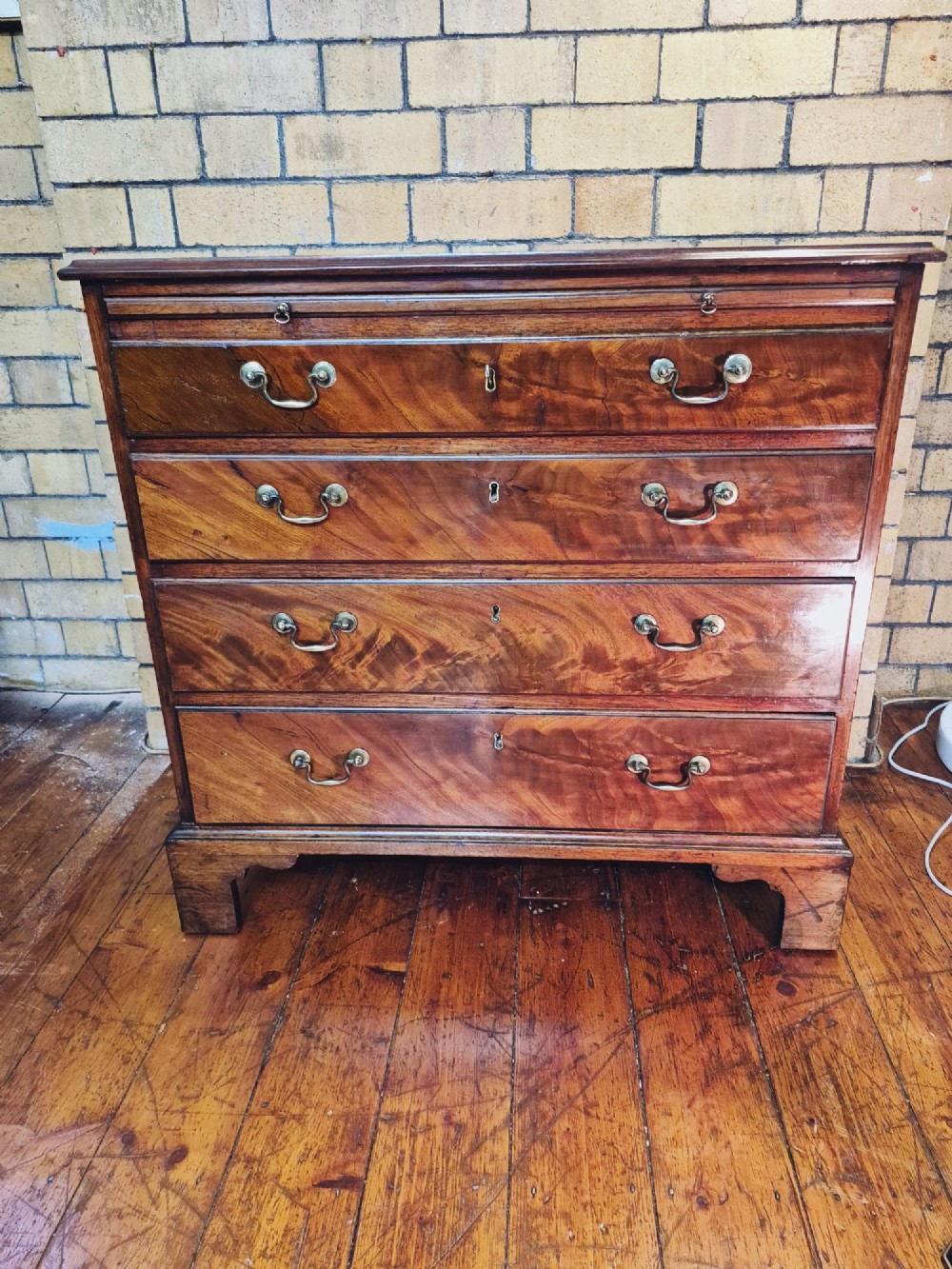 A Georgian mahogany bachelors chest featuring a top brushing slide, four graduated drawers with flame mahogany veneers, brass swan-neck handles, and bracket feet. View V