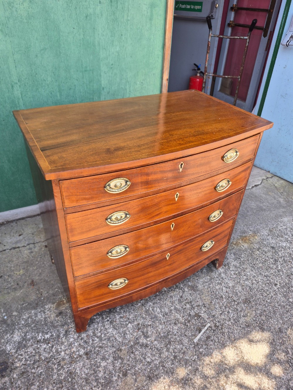 A Regency period mahogany bowfront chest featuring four graduated drawers with oval brass handles and splayed bracket feet. View V