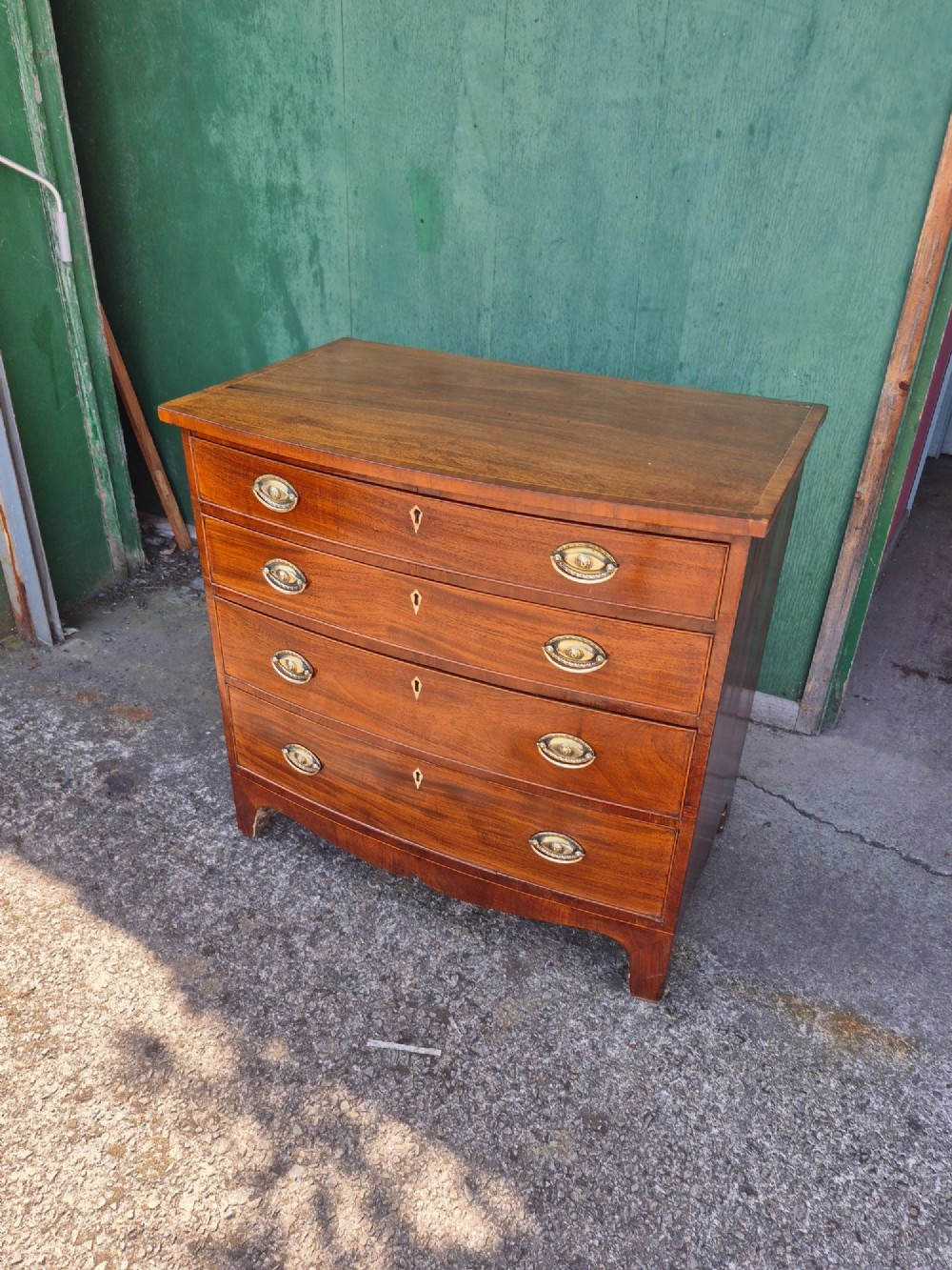 A Regency period mahogany bowfront chest featuring four graduated drawers with oval brass handles and splayed bracket feet. View VI