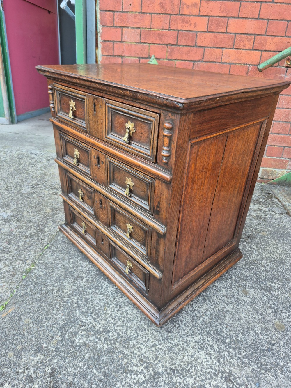 A small antique oak chest featuring four drawers with geometric moulding panels and brass drop handles. View II