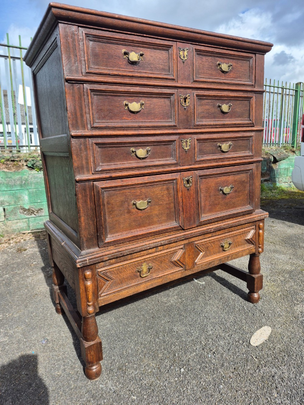 A 17th-century Charles II dark oak chest on stand featuring geometric panel mouldings, brass drop handles, and turned legs with connecting stretchers.