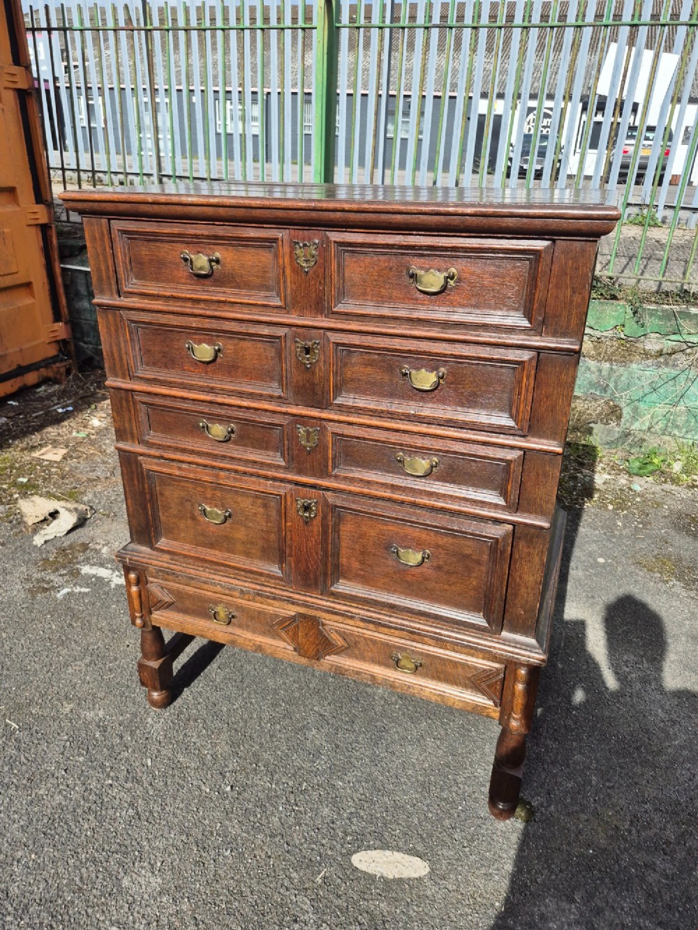 A 17th-century Charles II dark oak chest on stand featuring geometric panel mouldings, brass drop handles, and turned legs with connecting stretchers. View V