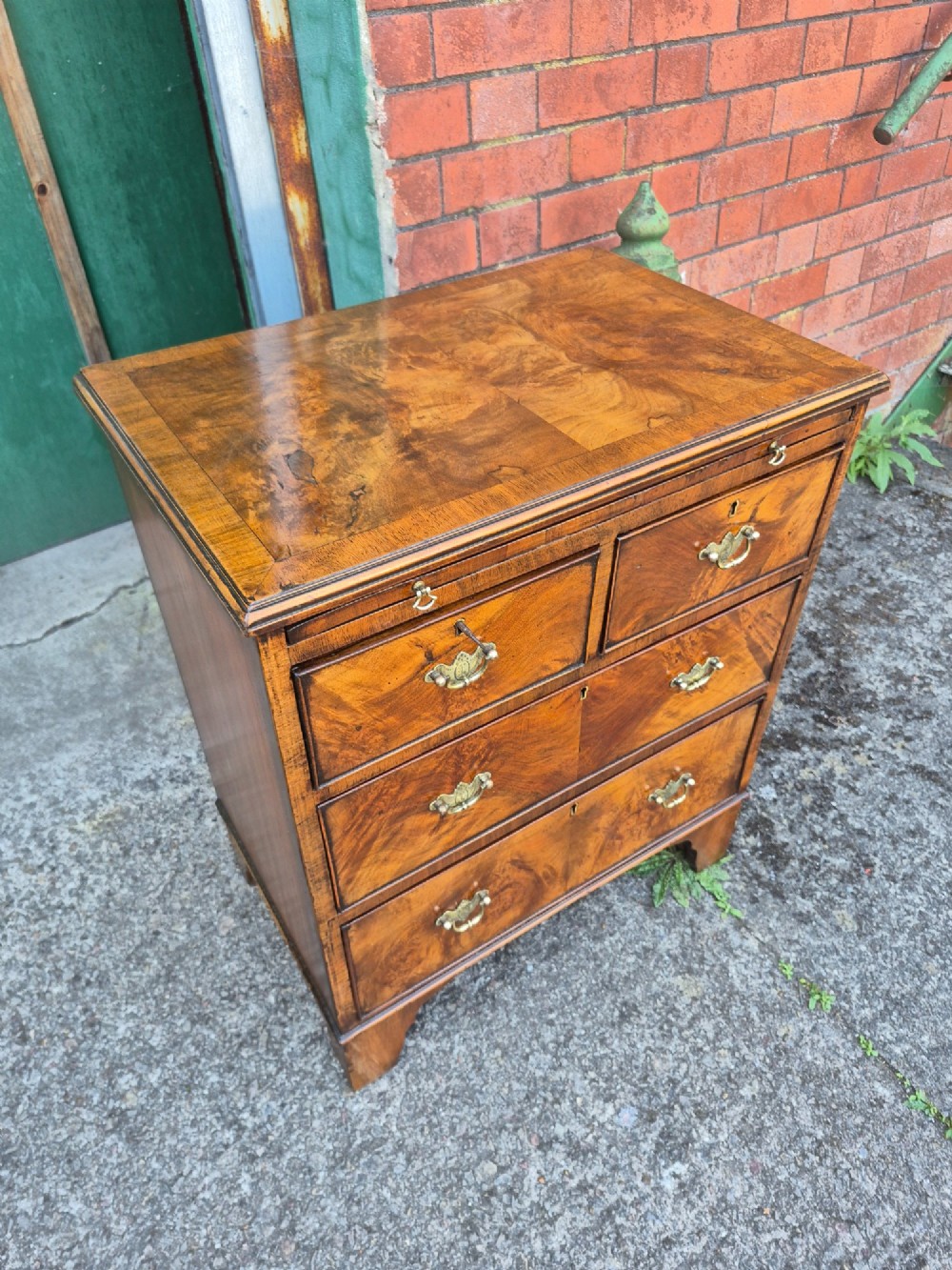 A small burr walnut bachelors chest featuring four drawers with brass handles, a brushing slide, and bracket feet. View VI