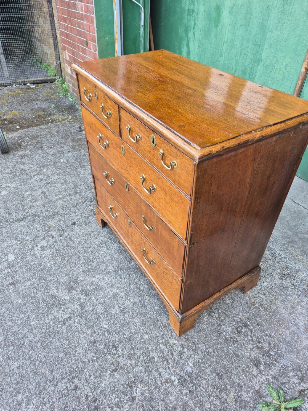 A George II golden oak chest of drawers featuring two short drawers over three long graduated drawers with brass swan-neck handles and bracket feet. View V