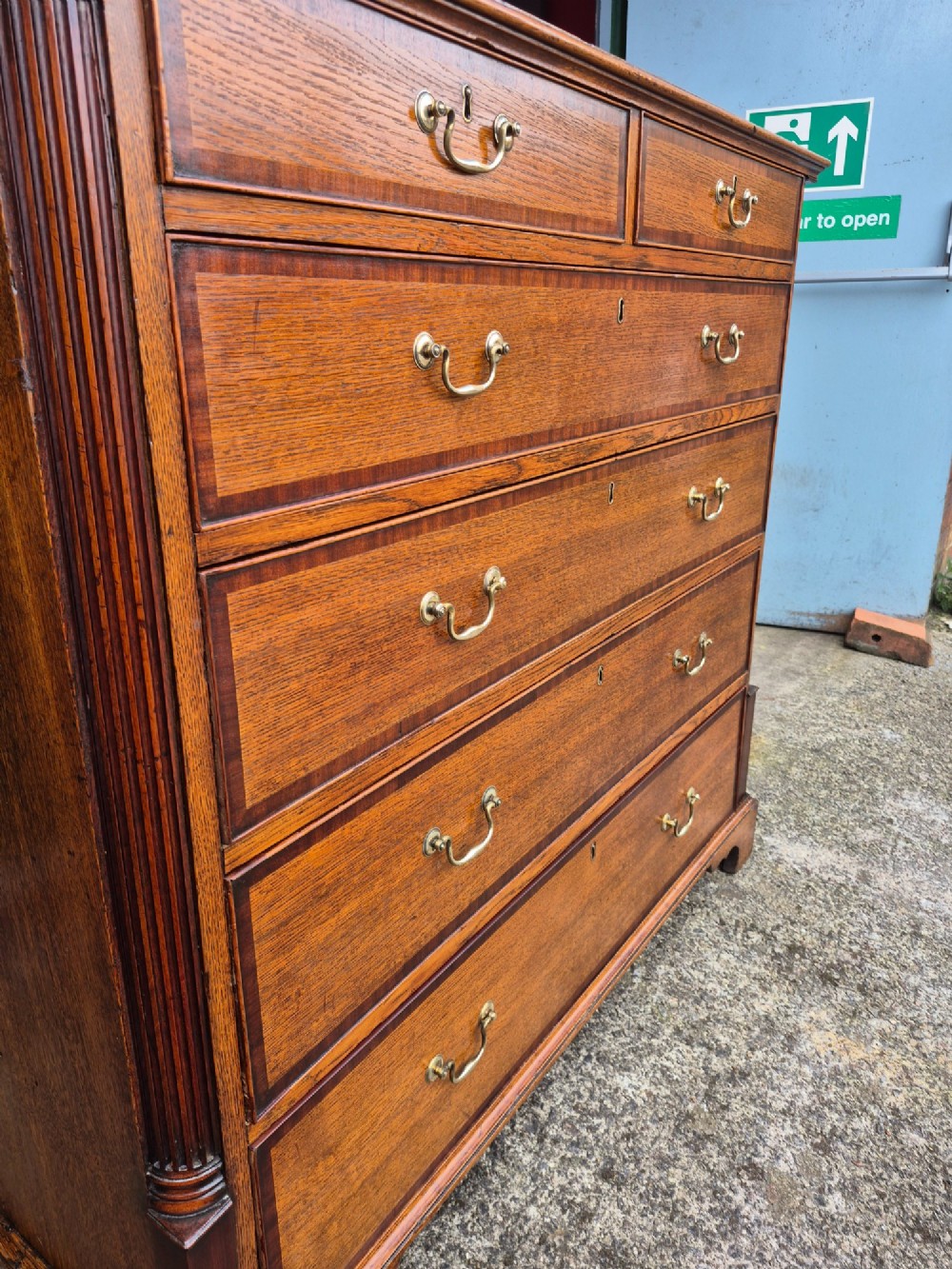 A golden oak Georgian chest of drawers featuring two short drawers over three long drawers with brass handles, reeded side columns, and bracket feet. View VI
