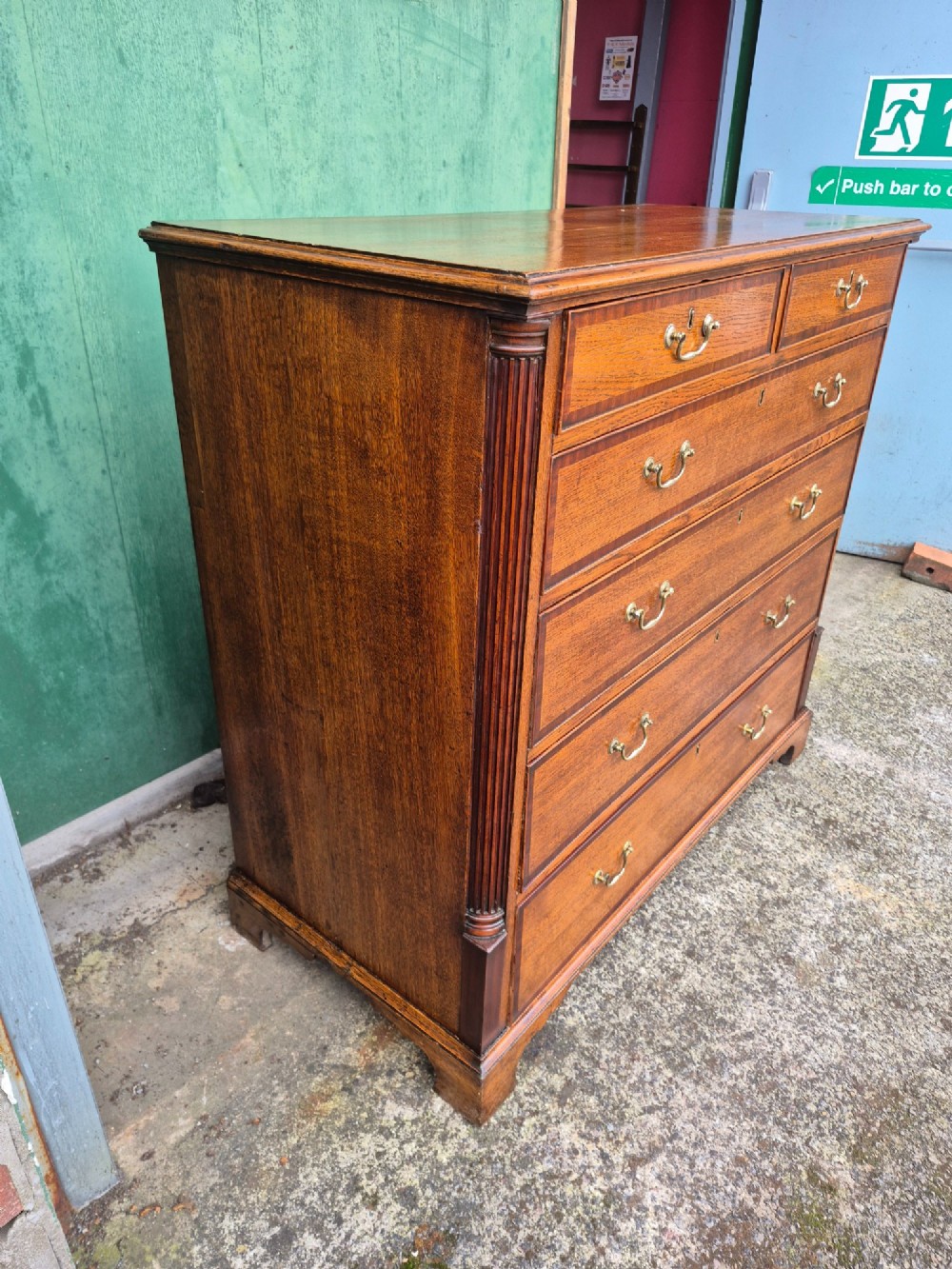 A golden oak Georgian chest of drawers featuring two short drawers over three long drawers with brass handles, reeded side columns, and bracket feet. View IX
