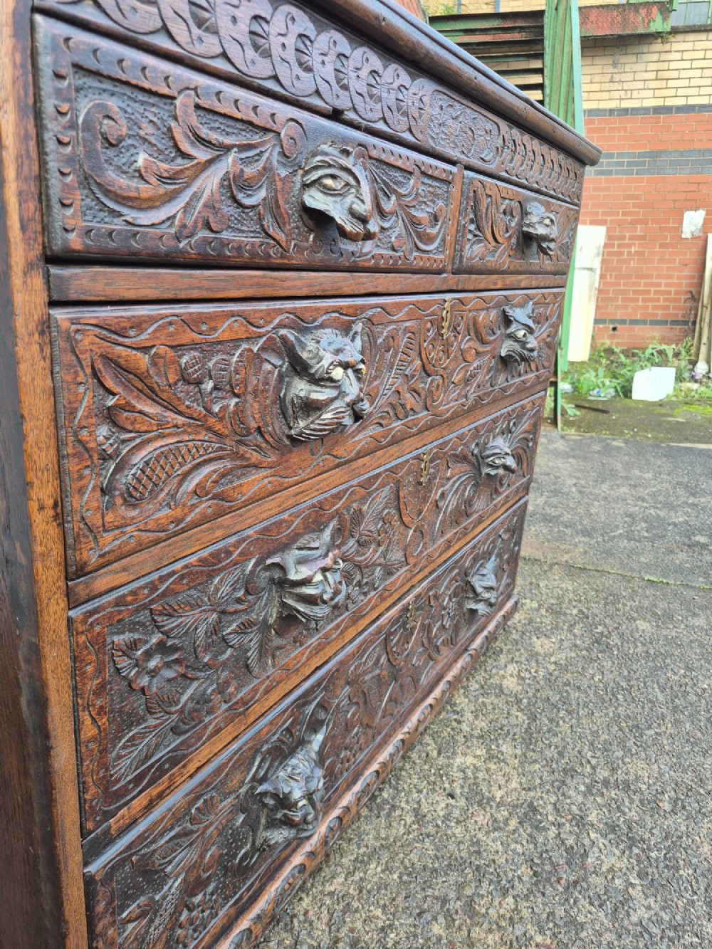 A nineteenth-century dark oak chest of drawers featuring hand-carved grotesque Green Man face masks and elaborate foliate scrollwork across the front. View V