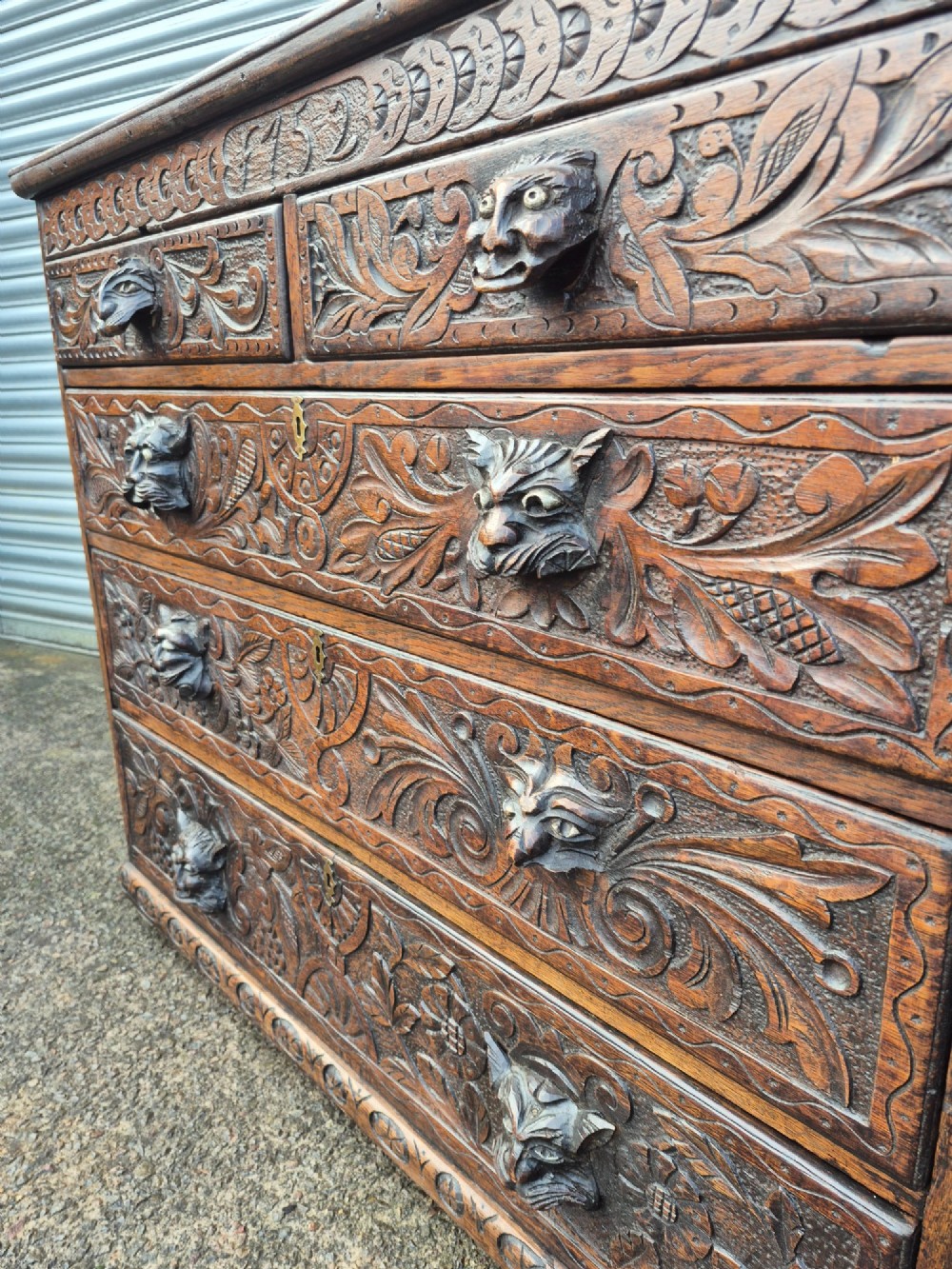 A nineteenth-century dark oak chest of drawers featuring hand-carved grotesque Green Man face masks and elaborate foliate scrollwork across the front. View VI