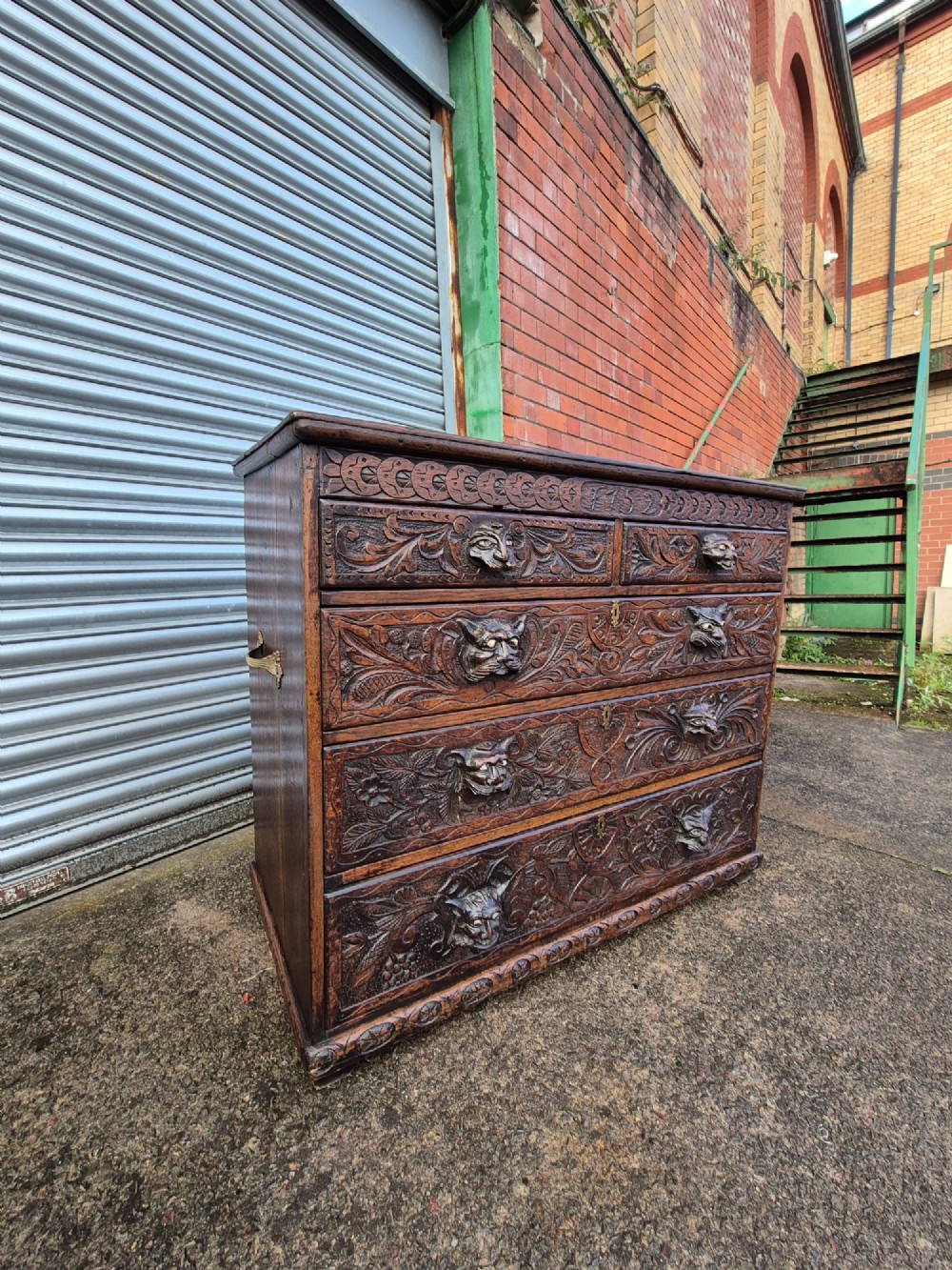 A nineteenth-century dark oak chest of drawers featuring hand-carved grotesque Green Man face masks and elaborate foliate scrollwork across the front. View XII