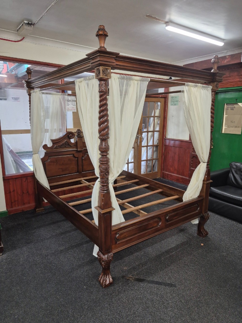 A dark hardwood queen-size four-poster bed frame featuring turned posts, a detailed carved headboard, and draped cream fabric. View X