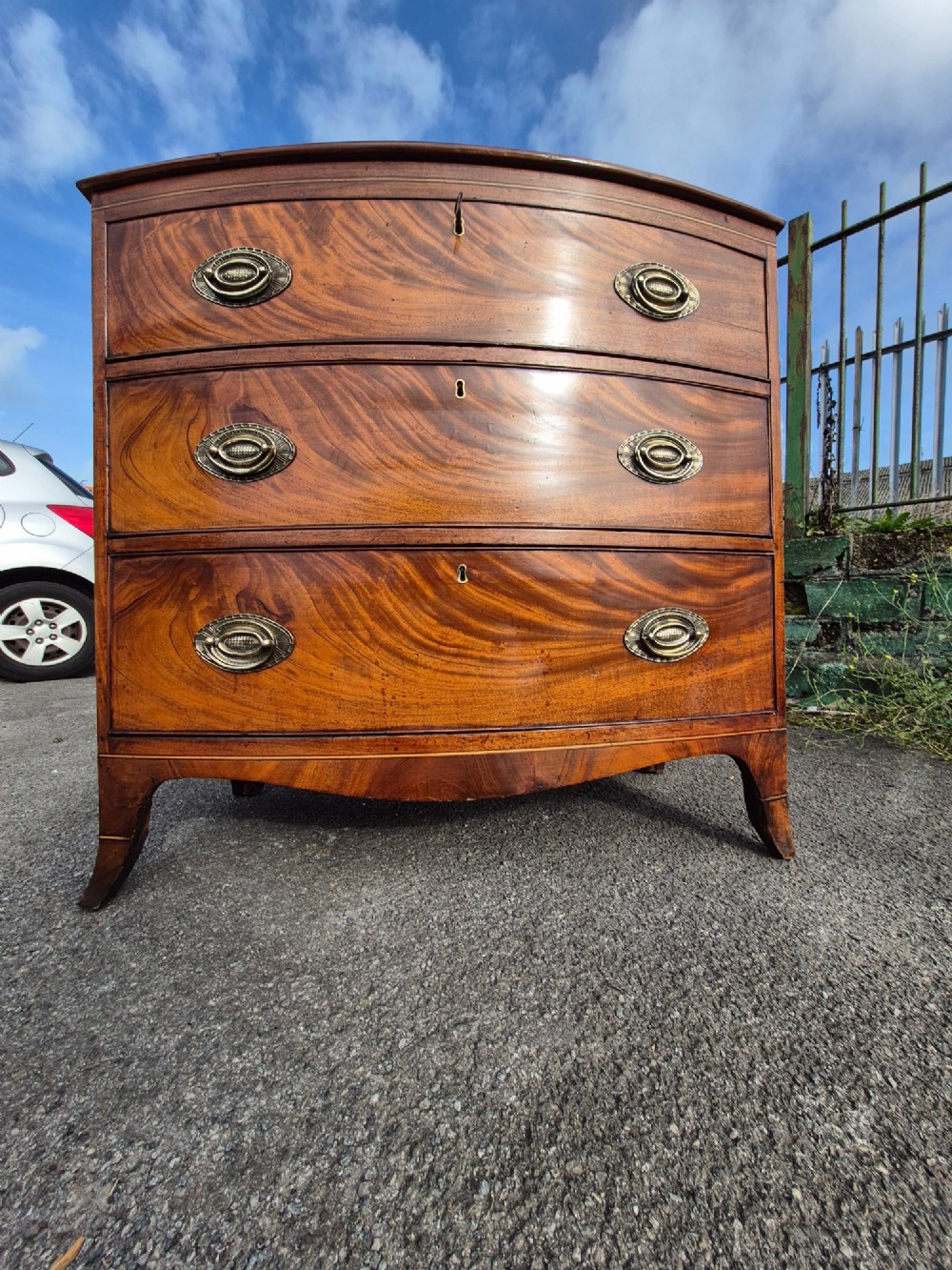 A Regency period bowfront chest of drawers crafted from flame mahogany veneer with three drawers and brass oval handles. View II