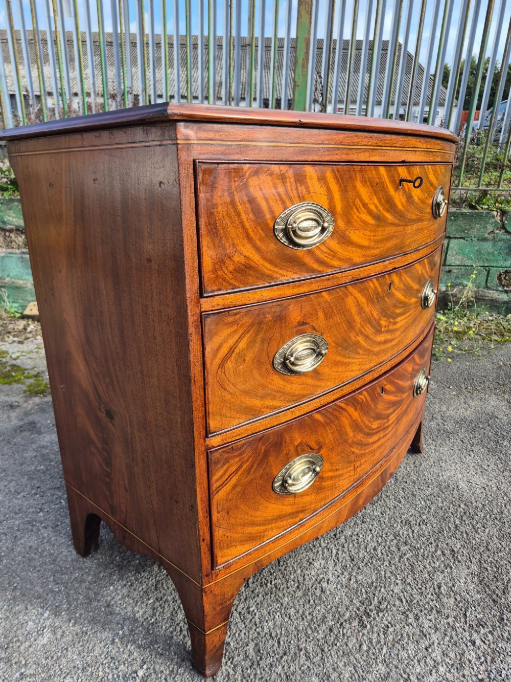A Regency period bowfront chest of drawers crafted from flame mahogany veneer with three drawers and brass oval handles. View X