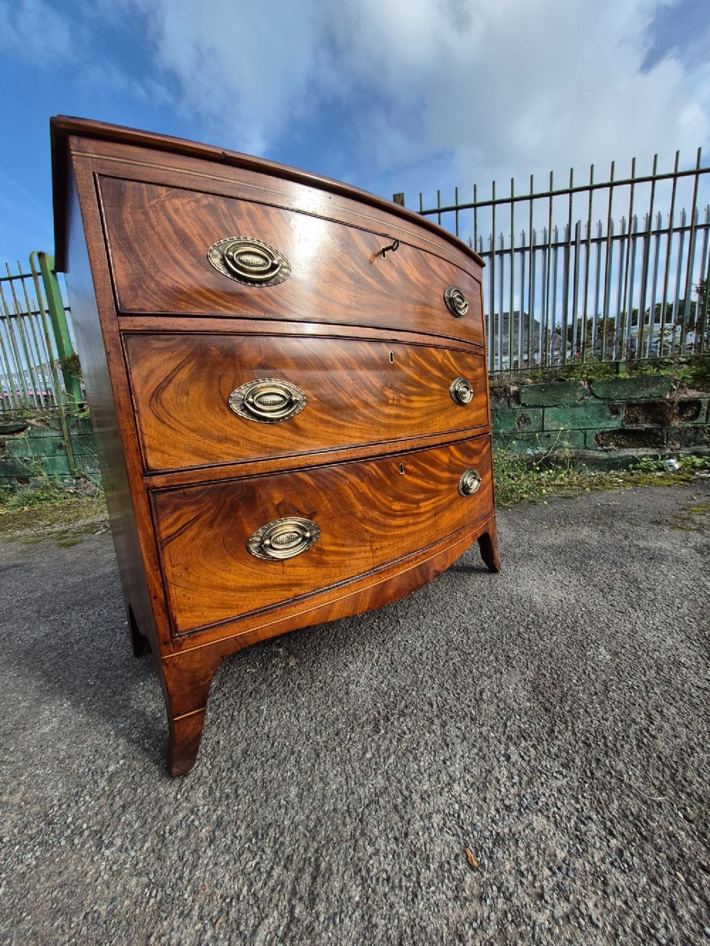 A Regency period bowfront chest of drawers crafted from flame mahogany veneer with three drawers and brass oval handles. View XI