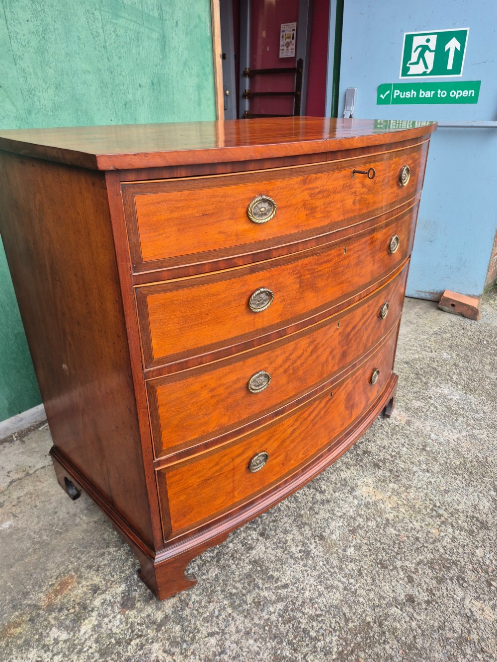 A Regency period satinwood bowfront chest of drawers featuring four graduated drawers with oval brass handles and bracket feet. View V
