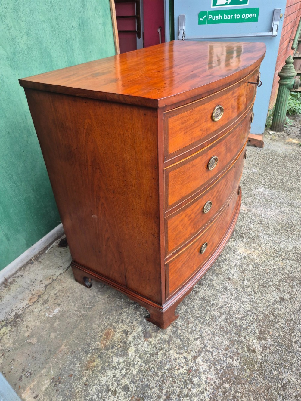 A Regency period satinwood bowfront chest of drawers featuring four graduated drawers with oval brass handles and bracket feet. View X