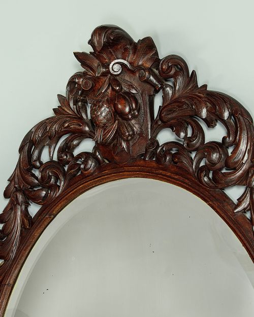 A late Victorian oval wall mirror featuring a carved dark oak frame with acanthus scrollwork and a crest motif enclosing a bevelled glass mirror plate. View III