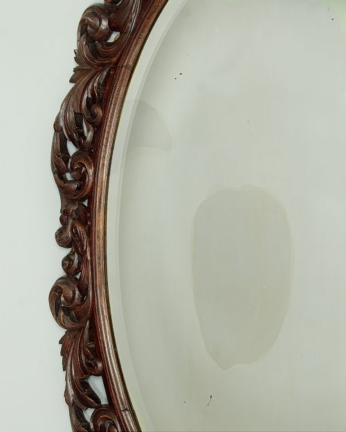 A late Victorian oval wall mirror featuring a carved dark oak frame with acanthus scrollwork and a crest motif enclosing a bevelled glass mirror plate. View V