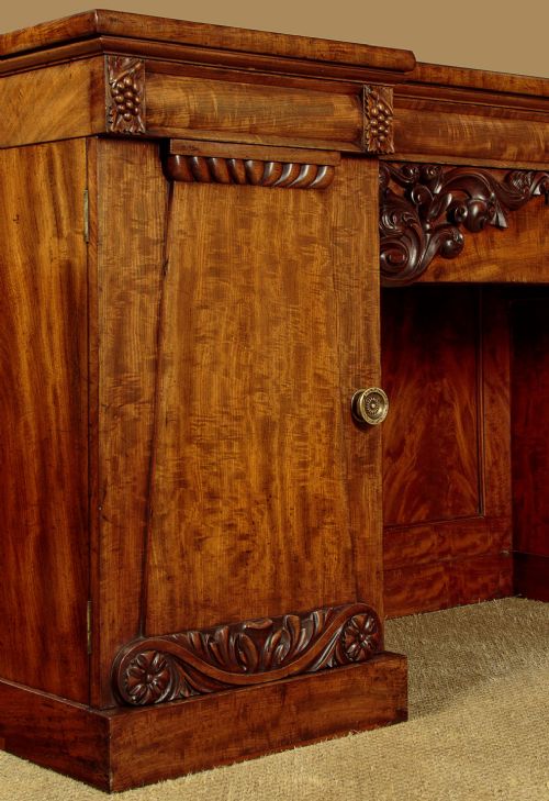 An early Victorian flame mahogany pedestal sideboard featuring carved scrollwork across the central apron and pedestal cupboards set on plinth bases. View IX