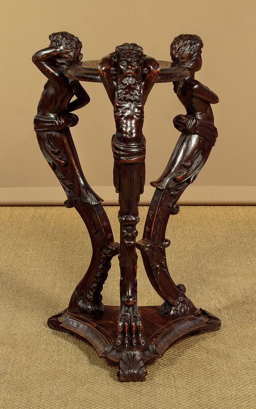 A dark carved wooden pedestal stand featuring three figurative sculpted supports on a tripartite base supporting a circular carved top. View II