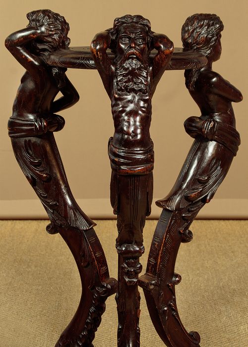 A dark carved wooden pedestal stand featuring three figurative sculpted supports on a tripartite base supporting a circular carved top. View V