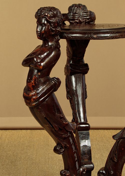 A dark carved wooden pedestal stand featuring three figurative sculpted supports on a tripartite base supporting a circular carved top. View IX