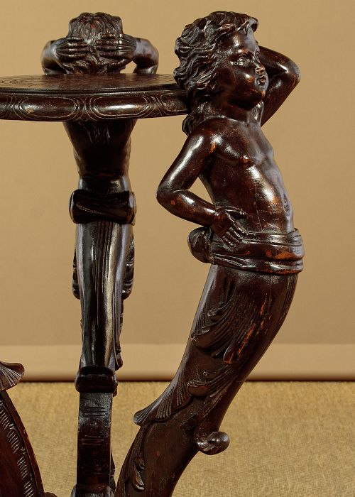 A dark carved wooden pedestal stand featuring three figurative sculpted supports on a tripartite base supporting a circular carved top. View X